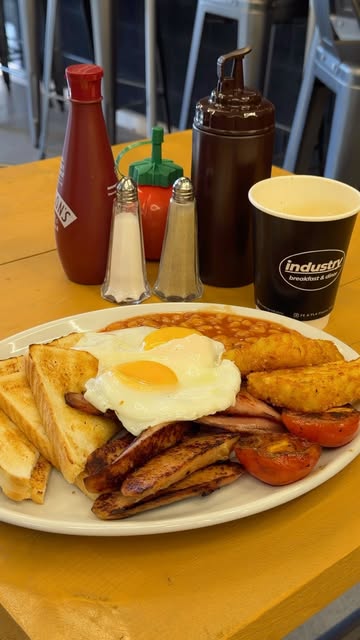Industry’s fry-up with eggs, bacon, sausages, hash browns, beans and toast, beside a branded cup